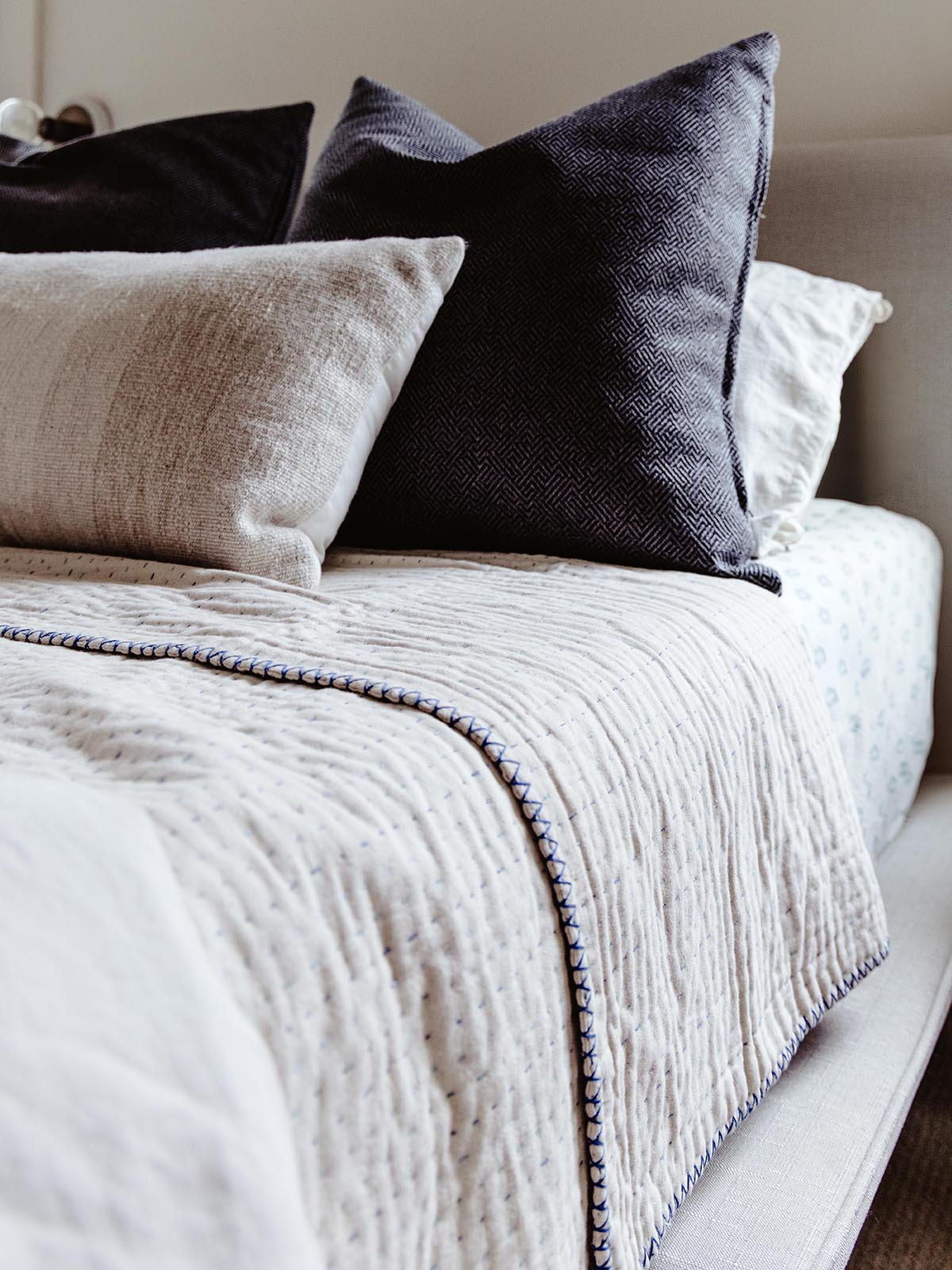 Close-up of a neatly made bed featuring a cream-colored quilt with blue stitching, complemented by dark and light gray accent pillows for a cozy and sophisticated look.