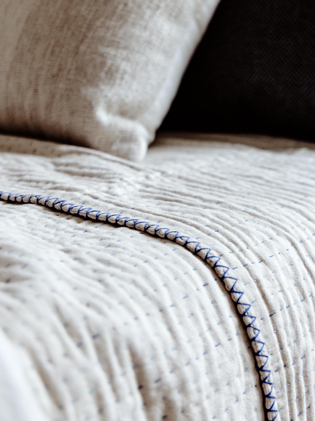 Detailed view of a cream-colored quilt with blue stitching on a bed, focusing on the texture and intricate hand-stitched edging, paired with a gray accent pillow.