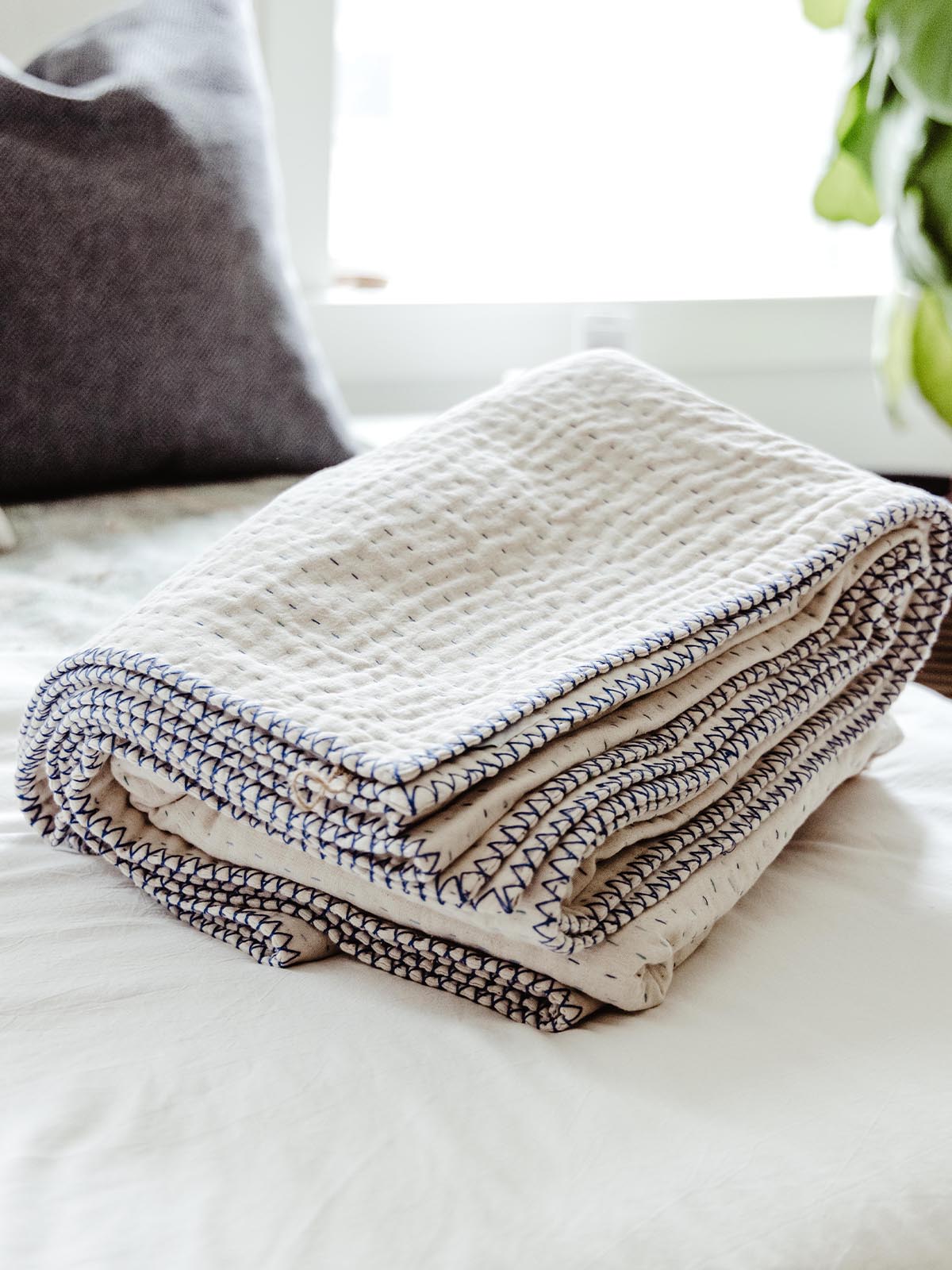 Folded cream-colored quilt with blue hand-stitched edges, resting on a bed, showcasing its soft texture and craftsmanship in a bright and airy bedroom setting.