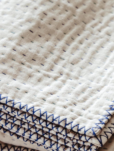 Close-up of a folded cream-colored quilt showcasing delicate hand-stitched blue and multicolored threads, highlighting the intricate craftsmanship and texture of the fabric.