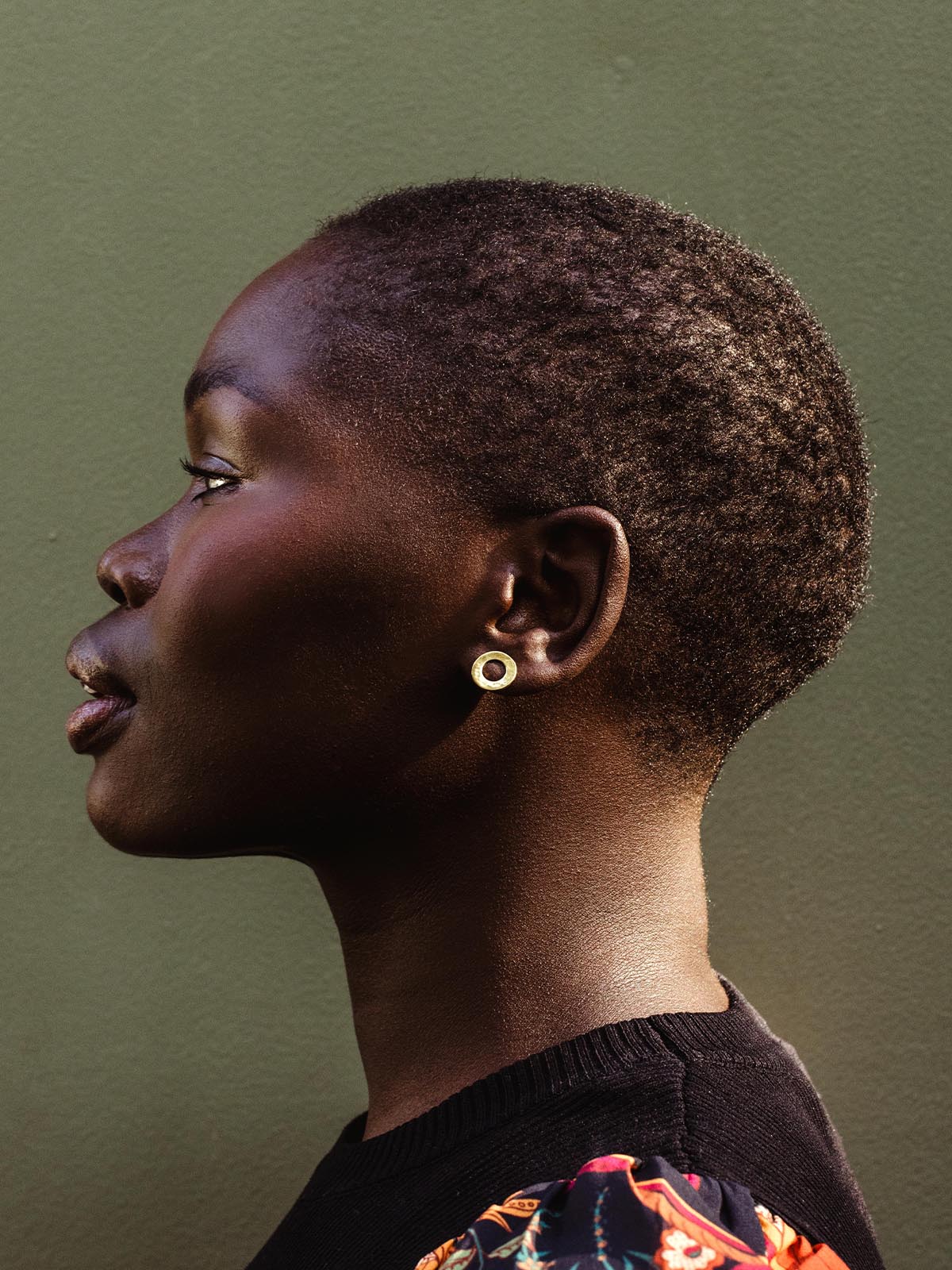 Side profile of a model with short hair wearing circular brass stud earrings against a muted green background.