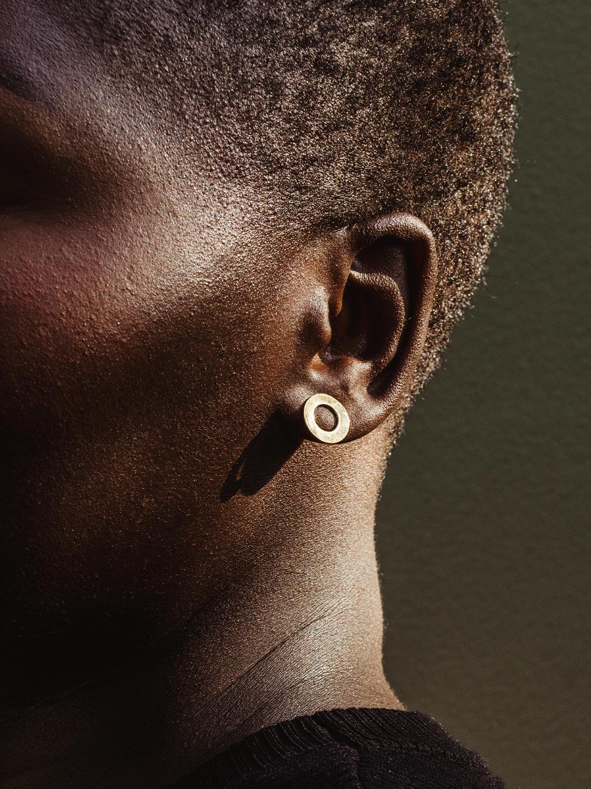 Close-up of a model's ear wearing a brass circular stud earring, capturing the texture and detail of the jewelry.