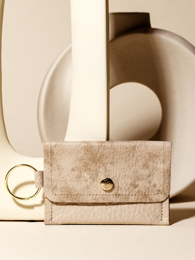 A stylish beige leather keychain wallet with a gold snap closure and keyring, featuring a minimalist design, displayed against modern ceramic decor for a sophisticated and functional accessory look.