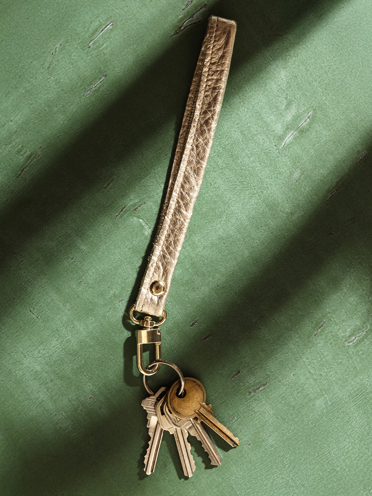 Platinum leather wristlet keychain with attached keys, featuring a sturdy gold clasp, displayed against a textured green background. A stylish and practical accessory for organizing keys with elegance.