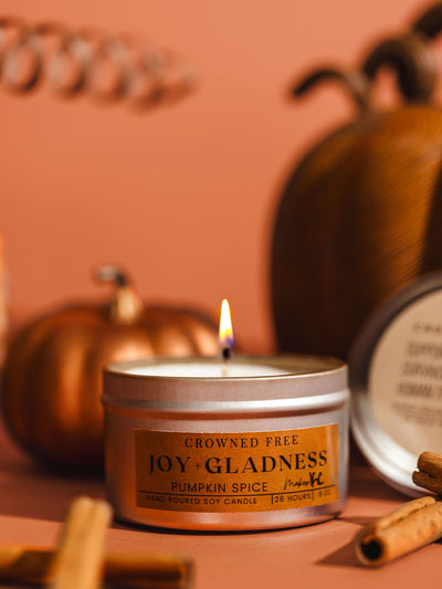 Lit pumpkin spice scented soy candle in a tin, labeled 'Joy + Gladness' by Crowned Free, surrounded by cinnamon sticks and decorative pumpkins, evoking a warm and cozy autumn atmosphere.