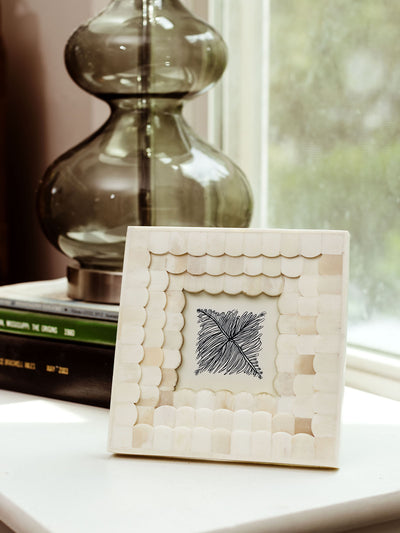 White scalloped frame made from bone, holding a black and white botanical illustration, placed on a white surface near a stack of books and a glass lamp, with natural light streaming in from a nearby window.