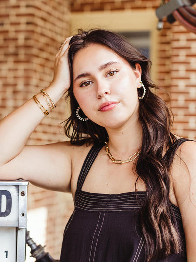 Close-up of a woman with long dark hair wearing silver hoop earrings and a gold chain necklace, standing against a brick wall with her hand resting on her head.