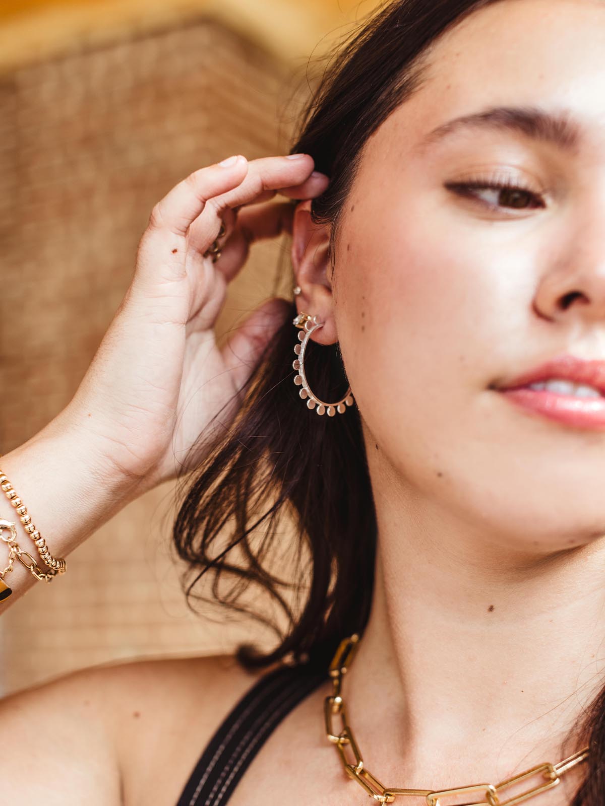 Close-up of a woman adjusting her silver hoop earring, showcasing layered bracelets and a gold chain necklace, with a blurred brick wall background.