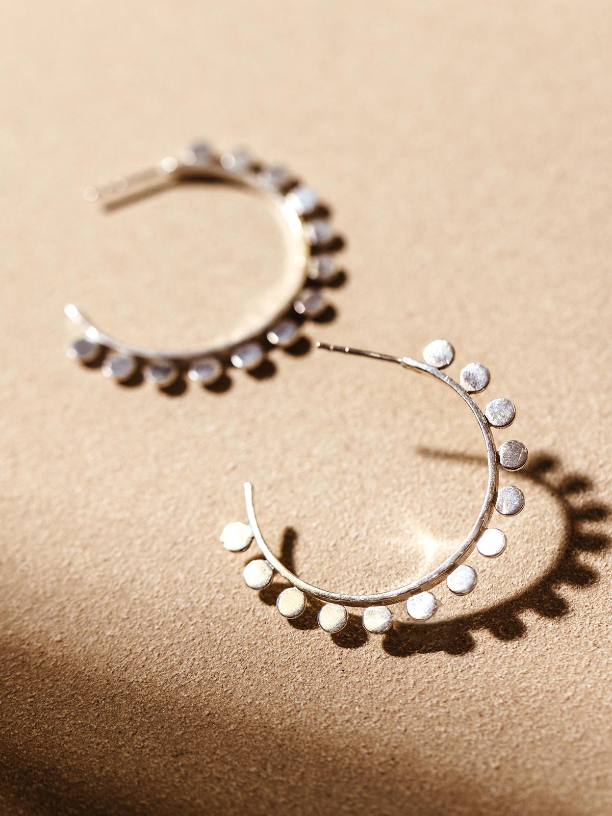 Close-up of a pair of textured silver hoop earrings with small circular details, displayed on a neutral surface with warm lighting.