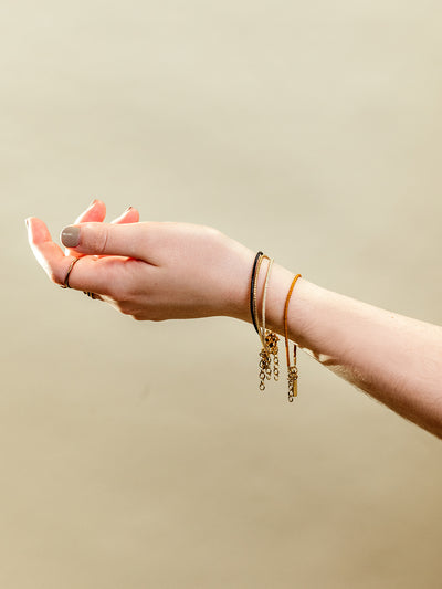 Close-up of an outstretched hand adorned with stackable gold rings and delicate beaded bracelets in black, mustard yellow, and gold tones, featuring gold chain accents, set against a neutral beige background to highlight minimalist handcrafted jewelry.