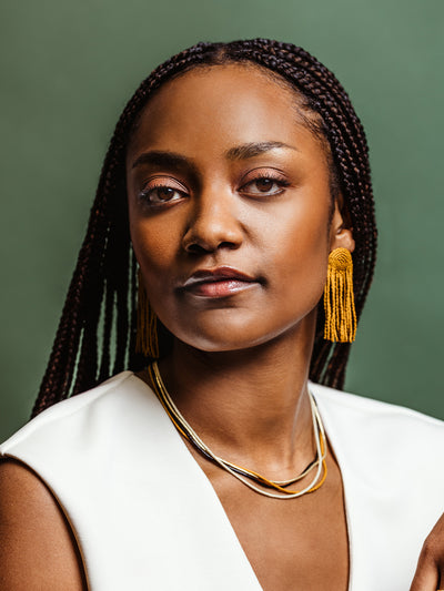 Close-up portrait of a woman wearing mustard yellow beaded tassel earrings and a layered gold and beaded necklace in black, ivory, and mustard tones, styled with a white outfit against a green background, showcasing elegant handcrafted jewelry.