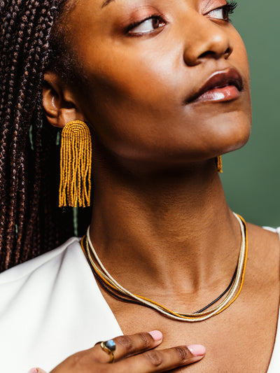 Detailed view of a woman wearing mustard yellow beaded tassel earrings and a layered beaded necklace in neutral tones, styled with a white top and a statement ring, emphasizing sustainable artisan jewelry.