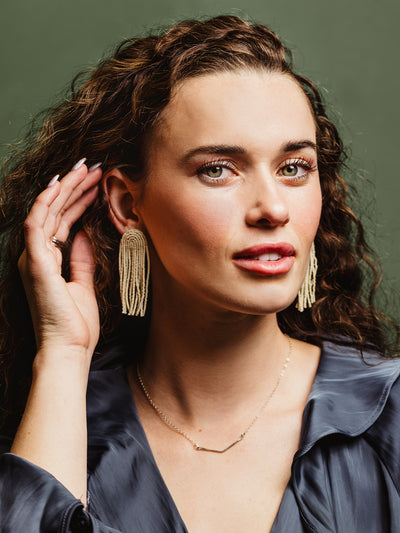 Portrait of a woman wearing cream-colored beaded tassel earrings and a dainty gold bar necklace, styled with a navy satin blouse, highlighting timeless and handcrafted jewelry.