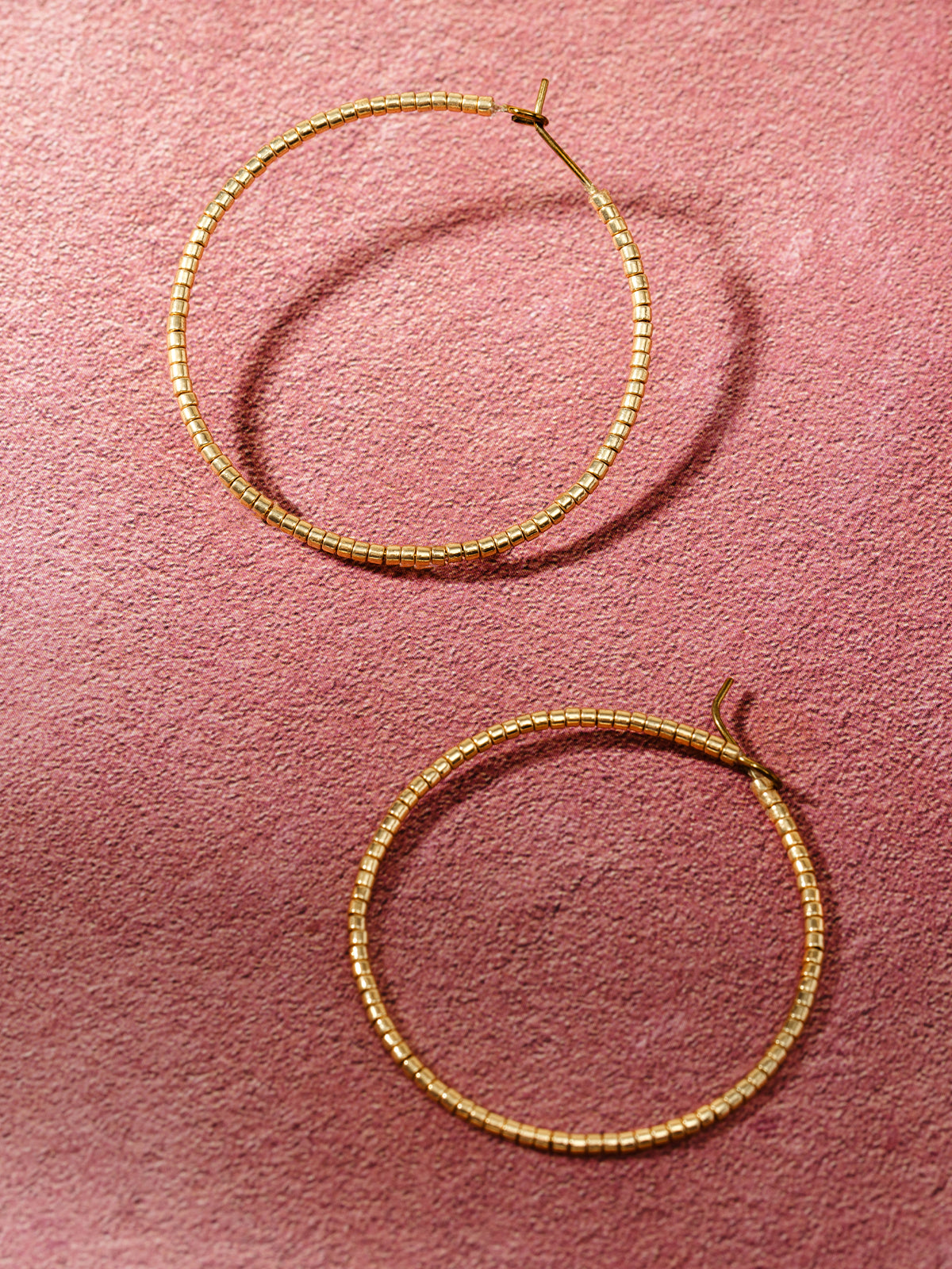 A pair of gold-toned beaded hoop earrings displayed on a soft pink textured background, showcasing their elegant and minimalistic design.