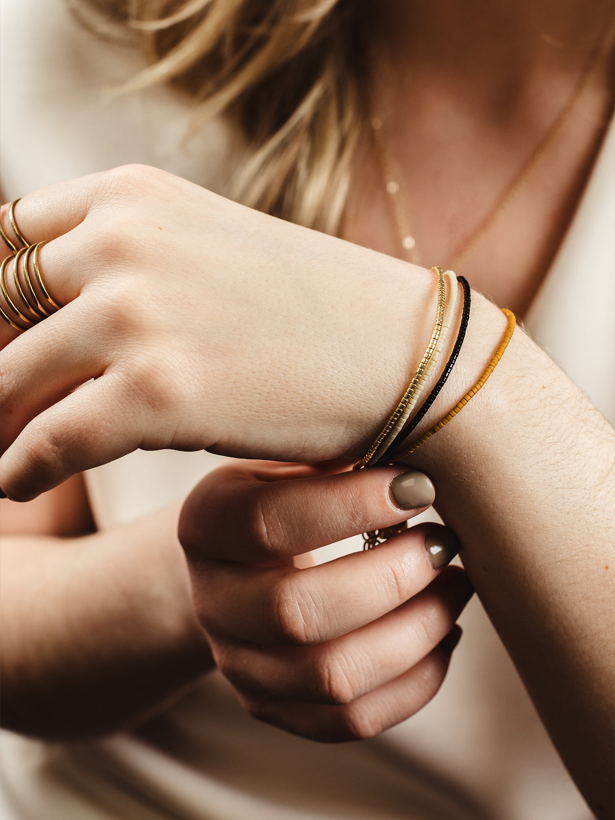 Close-up of a woman's wrist adorned with delicate beaded bracelets in gold, black, and mustard tones, paired with a gold multi-band ring, styled against a neutral outfit for a chic and modern look.