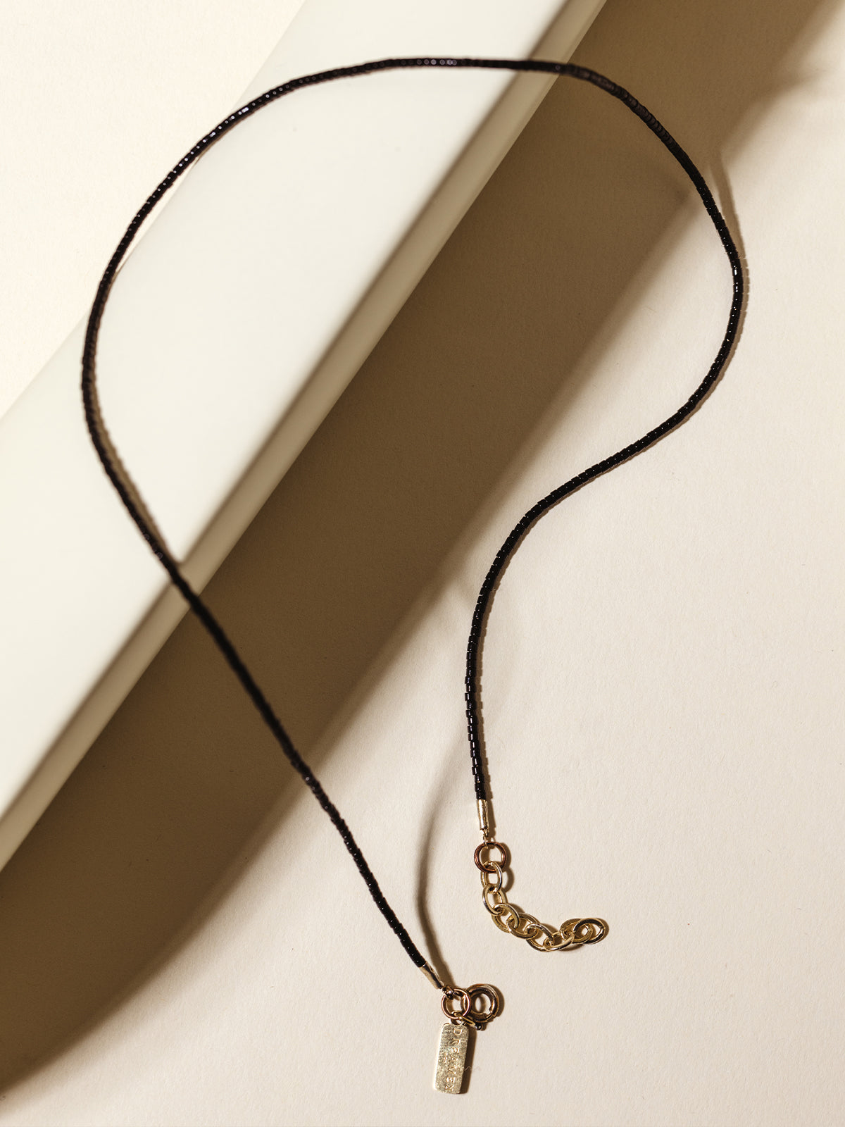  A black beaded necklace with a gold charm and extender chain, elegantly styled over a white ceramic structure for a bold and modern look.