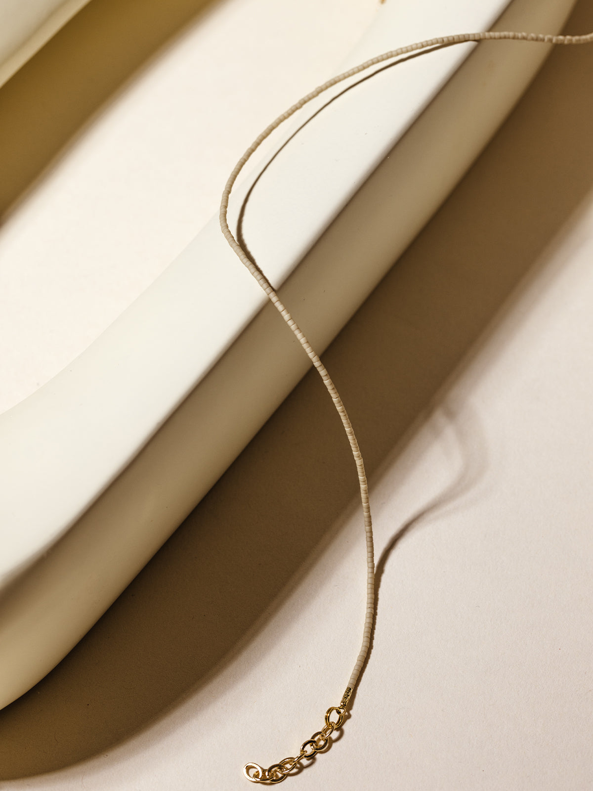 A neutral beige beaded necklace with a gold extender chain elegantly draped over a white ceramic form, showcasing its minimalist and timeless design