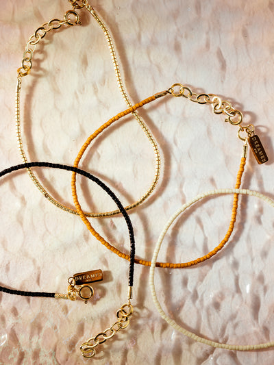 Delicate beaded necklaces in black, mustard yellow, ivory, and gold tones with gold chain accents and 'Dreamer' charm tags, displayed on a textured neutral background, showcasing artisan-crafted minimalist jewelry.