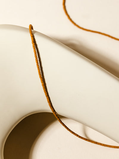 A mustard-hued beaded necklace with gold chain details displayed over a soft beige ceramic form, showcasing its earthy and warm tones.