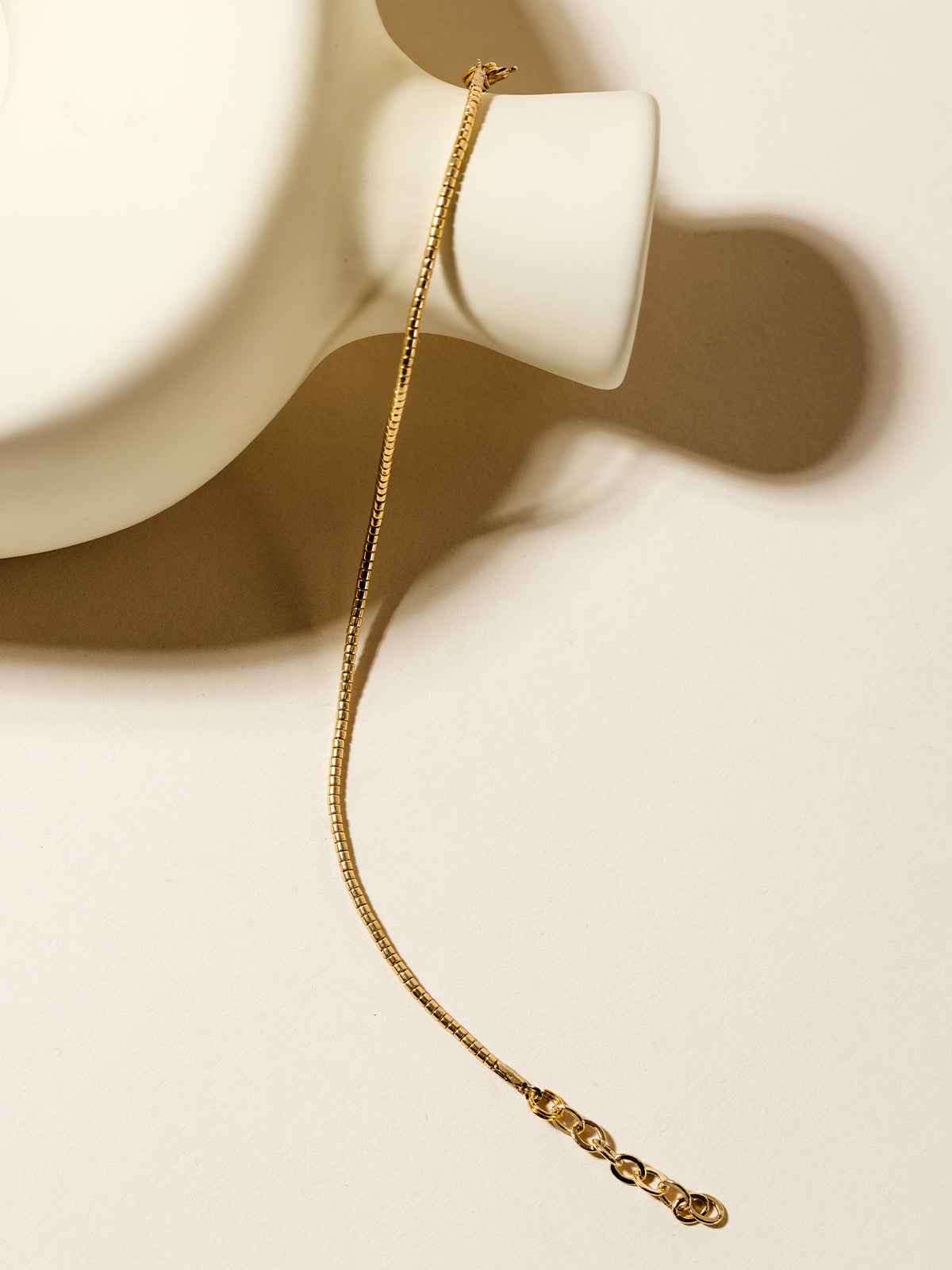 A delicate gold beaded bracelet draped over a modern white ceramic vase, highlighting its minimalist and timeless elegance against a neutral background.