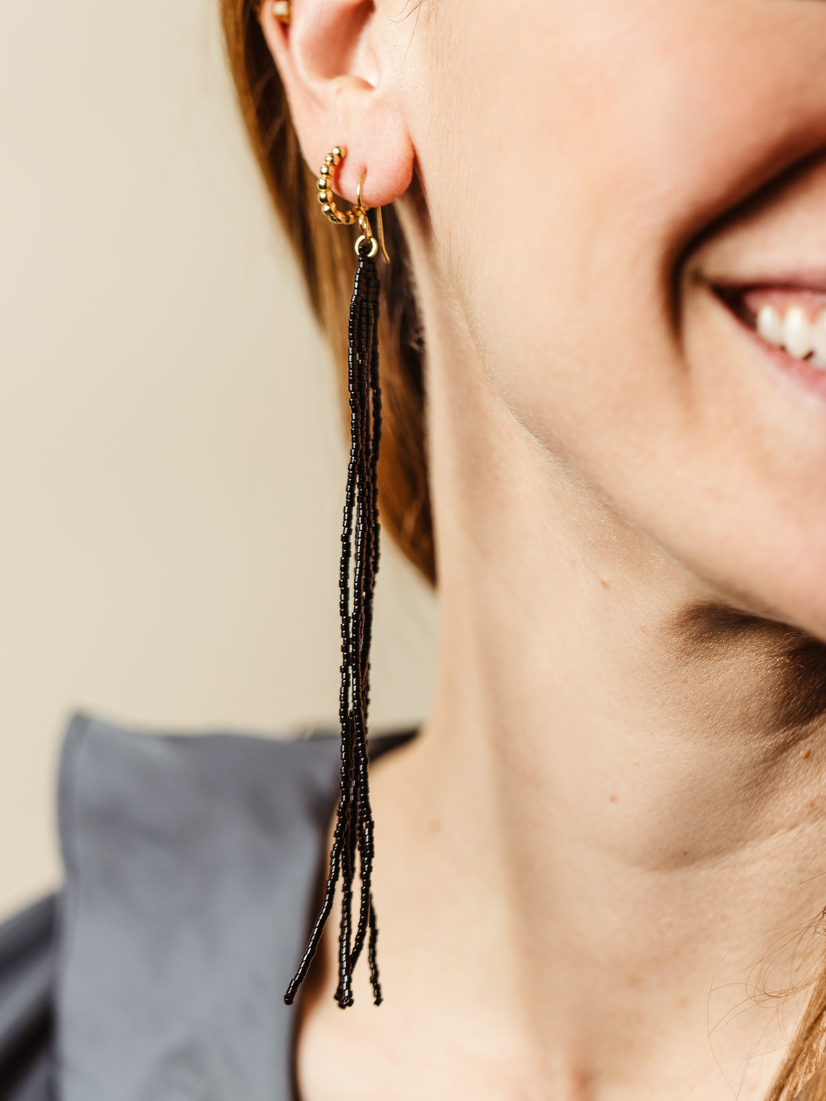 Smiling woman wearing black beaded tassel earrings with gold hoops, adding a touch of elegance and sophistication to her modern outfit.