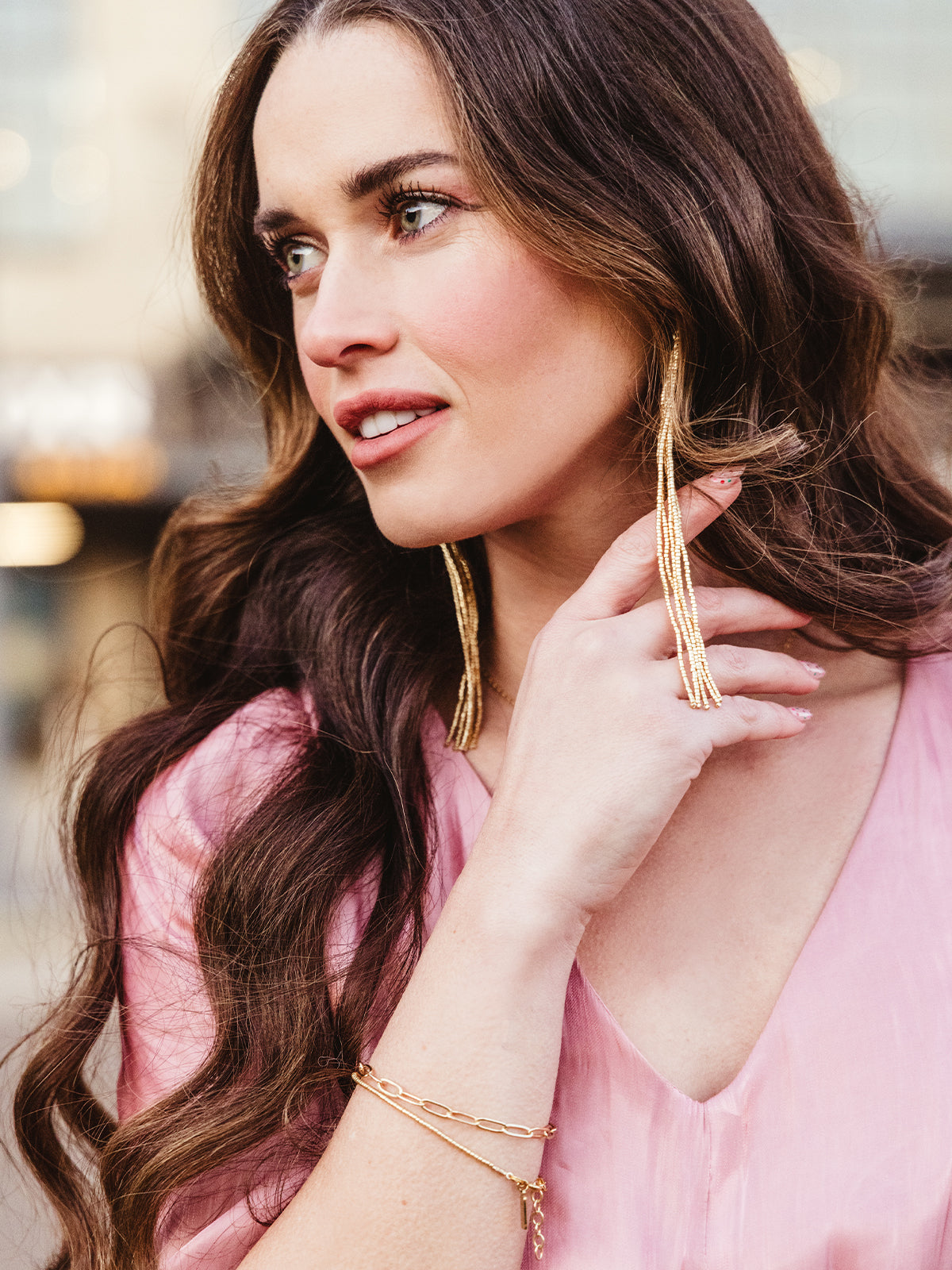 Close-up of a woman wearing cascading gold tassel earrings, a delicate gold bracelet, and a statement ring, styled with a soft pink blouse, showcasing elegant handcrafted jewelry for a sophisticated look.