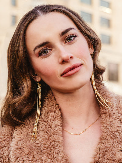 Portrait of a woman wearing long gold tassel earrings and a minimalist gold necklace, styled with a cozy tan faux fur coat, highlighting chic and handcrafted jewelry perfect for fall and winter fashion.