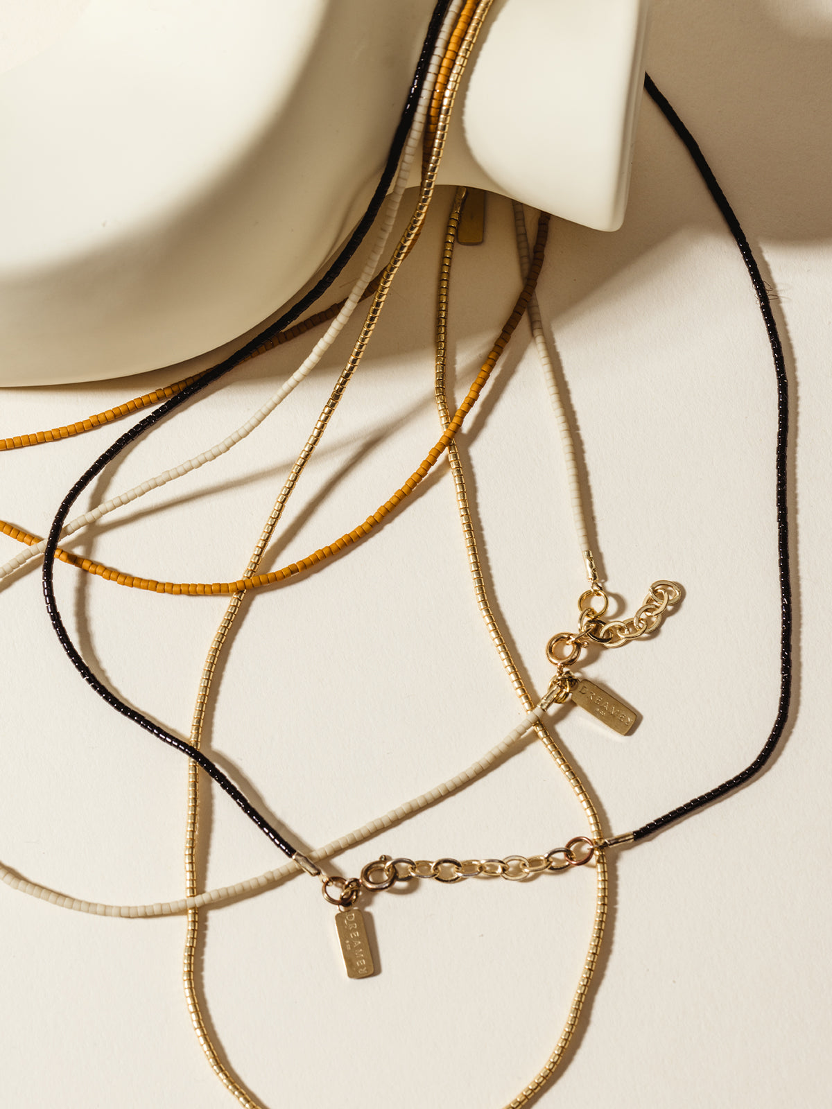 Close-up of delicate beaded necklaces in black, mustard yellow, ivory, and gold tones with gold chain clasps and 'Dreamer' tags, styled on a cream background, showcasing sustainable and handmade jewelry.