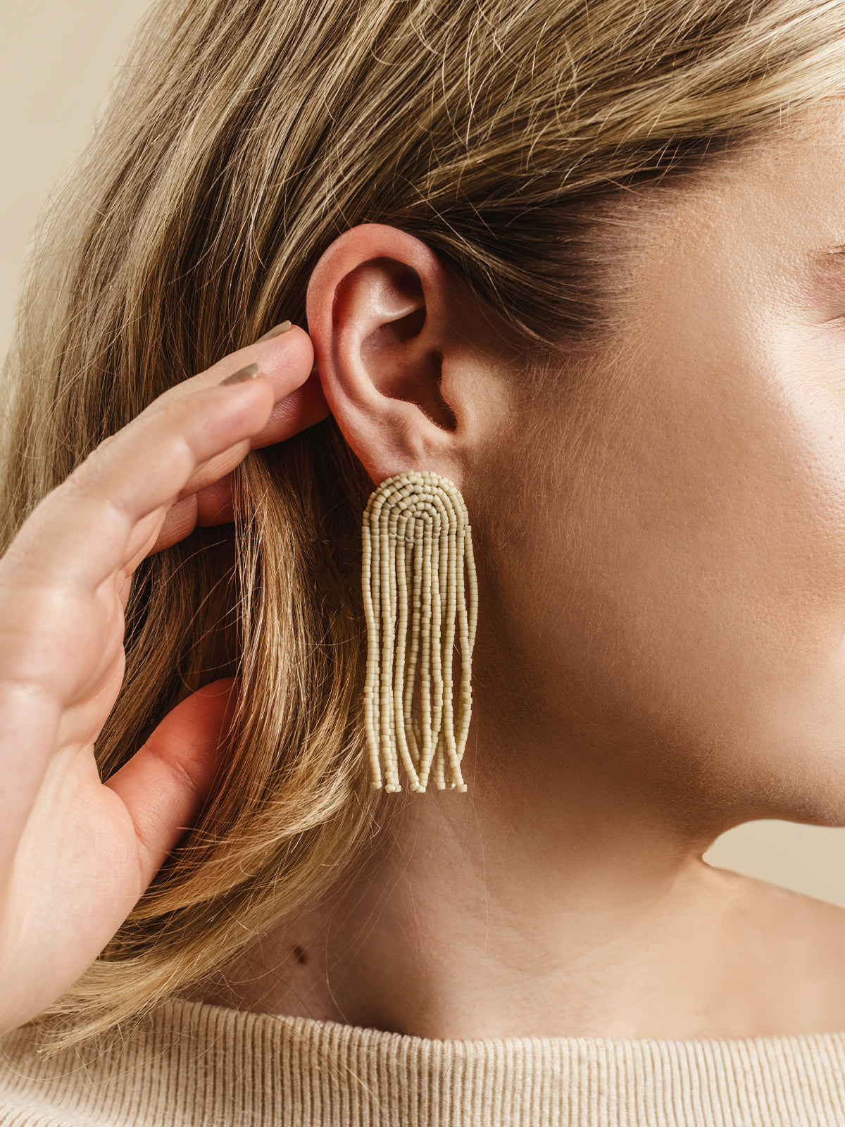 Close-up of a woman wearing handcrafted cream beaded tassel earrings, styled with blonde hair and a beige ribbed sweater, highlighting minimalist and elegant artisan jewelry for a timeless look.