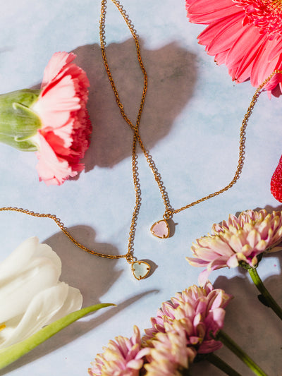 Two gold pendant necklaces, one featuring a soft pink gemstone and the other a pale green gemstone, displayed among pink and white flowers on a soft blue background. The floral arrangement enhances the delicate and feminine aesthetic of the jewelry.
