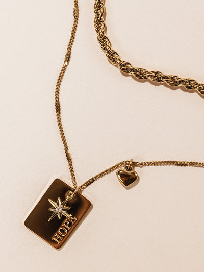 Statement gold necklace featuring two chains and rectangular pendant with "hope" etched into pendant. Also featuring a heat and star charm. Photographed on cream background. 