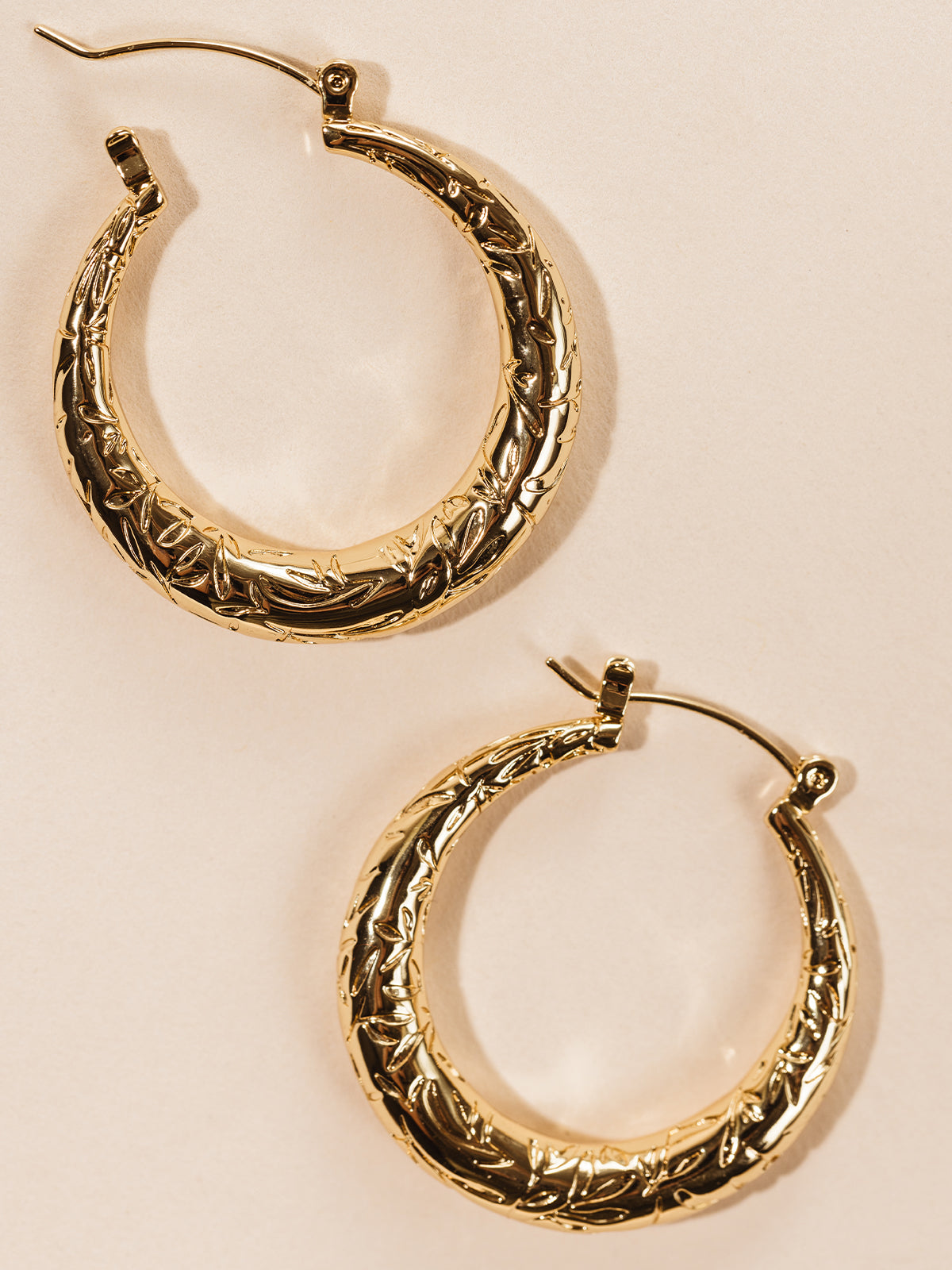 Gold hoop earrings with engraved leaf design on cream background. 