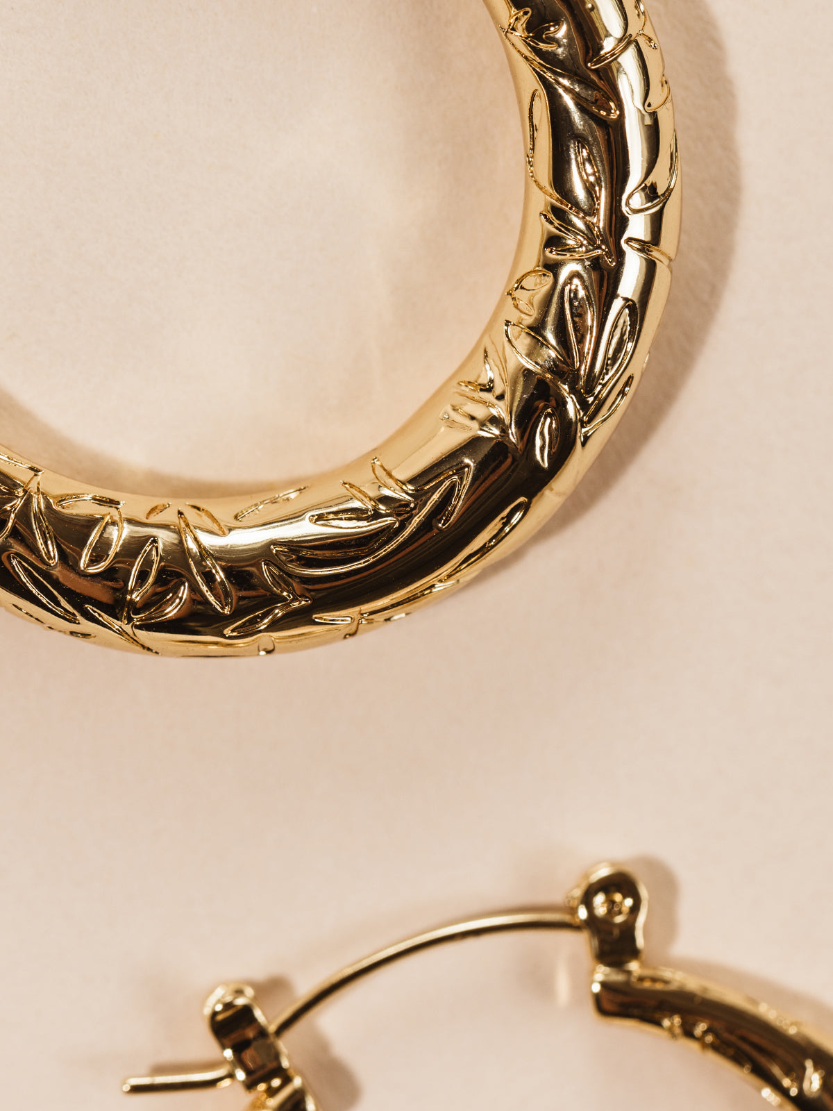 Gold hoop earrings with engraved leaf design on cream background. 