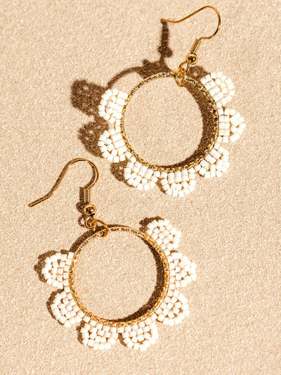 Gold hoop earrings with white beaded flowers arranged around the circle, creating a delicate and floral-inspired design. The earrings are set against a textured beige surface, highlighting their elegant craftsmanship.