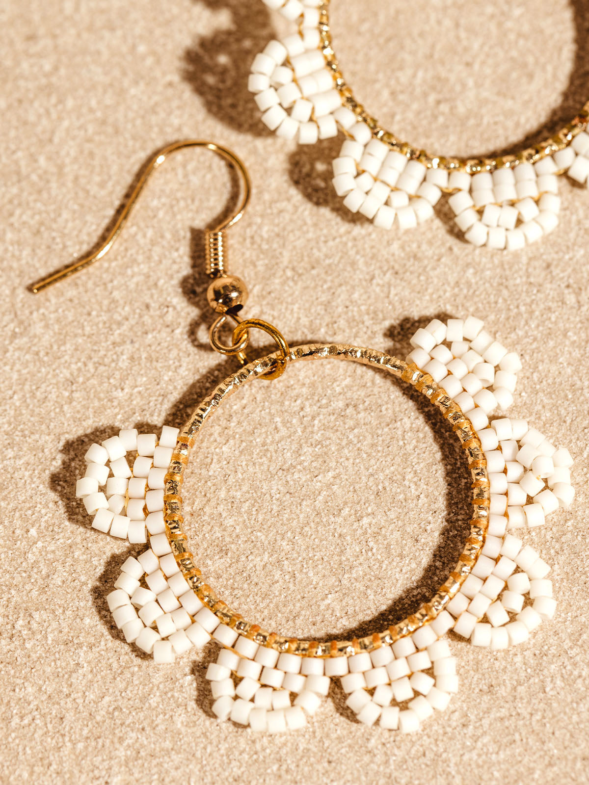 Close-up of gold hoop earrings featuring white beaded flowers, arranged to form a delicate and feminine design. The earrings are displayed on a neutral background, emphasizing their intricate and elegant details.