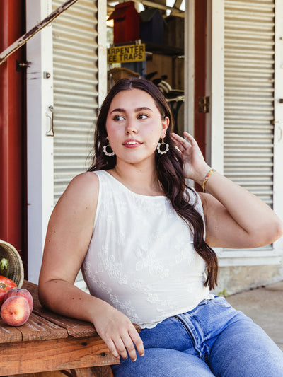 A young woman sits on a wooden bench outside a rustic building, wearing a white embroidered sleeveless top and blue jeans. She touches her long dark hair with one hand while the other rests on the bench near a basket of peaches and tomatoes. The woman is accessorized with large, circular beaded earrings and a colorful bracelet, adding a touch of elegance to the relaxed, rural setting.