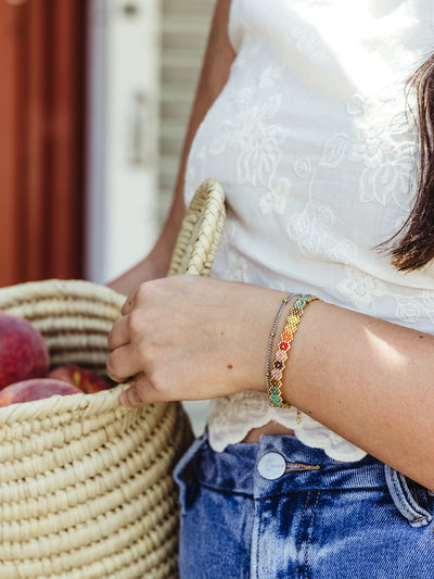 A close-up of a woman's hand holding a woven basket filled with peaches. She is wearing a white embroidered top and blue jeans. On her wrist, she wears a colorful beaded bracelet with a geometric floral pattern, adding a vibrant touch to the casual, rustic scene.