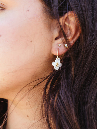 Close-up of a woman's ear wearing a small gold and white beaded earring with a delicate geometric design. The earring dangles just below her earlobe, complemented by two small stud earrings in her second and third piercings. Strands of her dark hair frame the side of her face, emphasizing the intricate details of the jewelry.