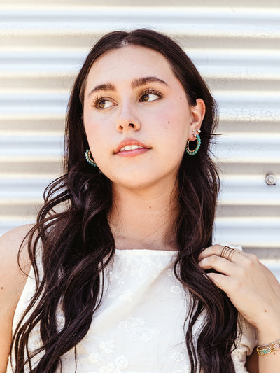 A young woman with long dark hair poses against a corrugated metal wall. She is wearing a white top with delicate floral embroidery and turquoise beaded hoop earrings. One hand gently touches her hair while the other rests near her collarbone, displaying several gold rings. The setting highlights her natural beauty and the handcrafted jewelry she wears.