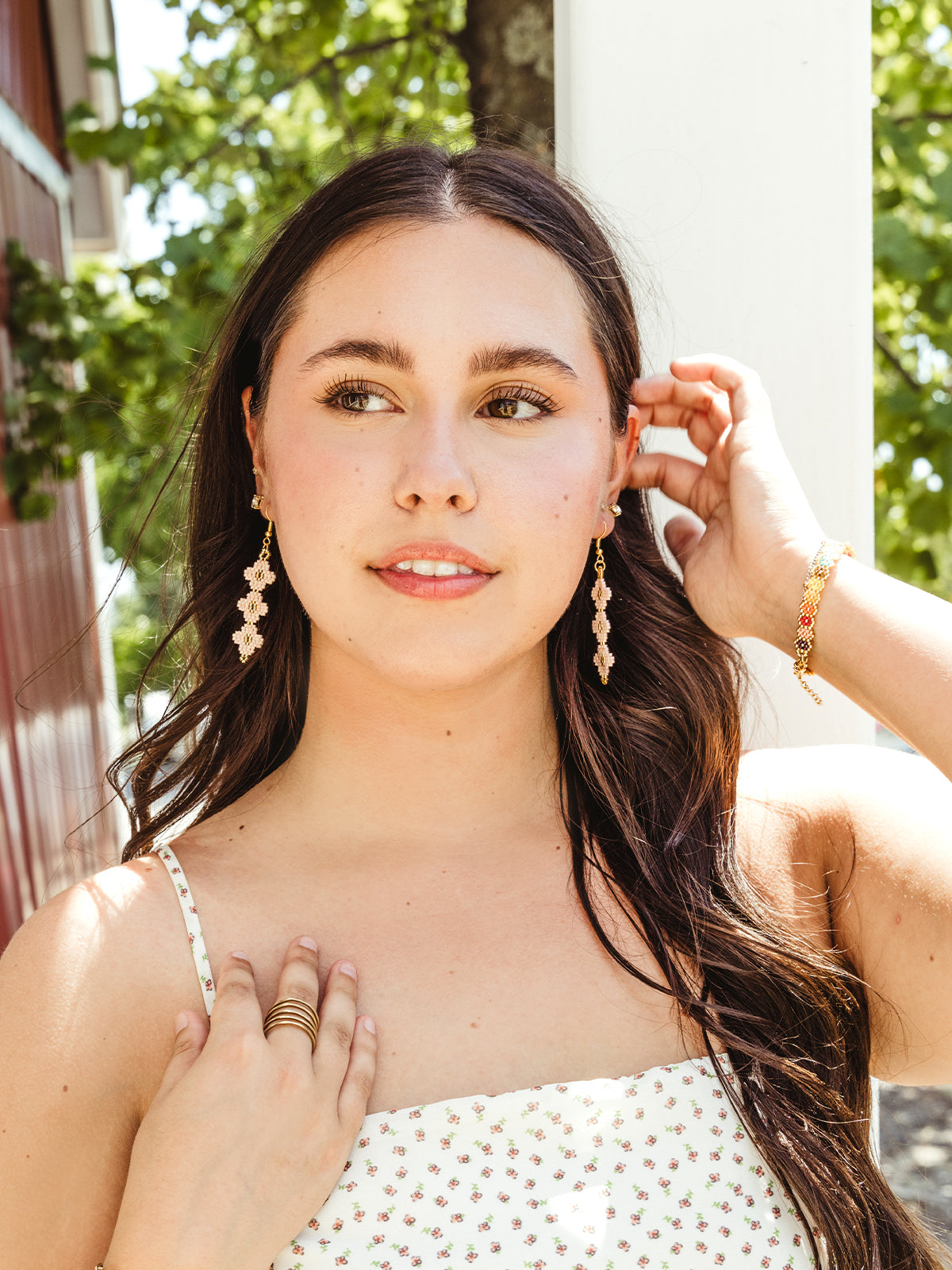 A young woman with long dark hair stands outdoors, wearing a white floral-patterned dress. She touches her hair with one hand and her collarbone with the other, showing off a pair of intricate beaded earrings in a soft pink and gold color. The background features a sunlit, leafy tree and a red barn wall.