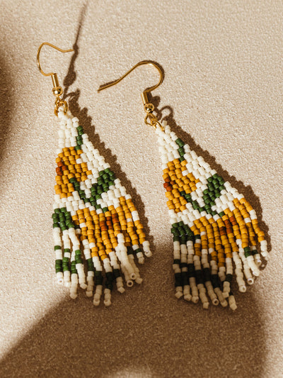 Beaded earrings with an intricate sunflower design, featuring yellow, green, and white beads. The earrings are laid out on a textured beige surface, showcasing their vibrant colors and detailed craftsmanship.