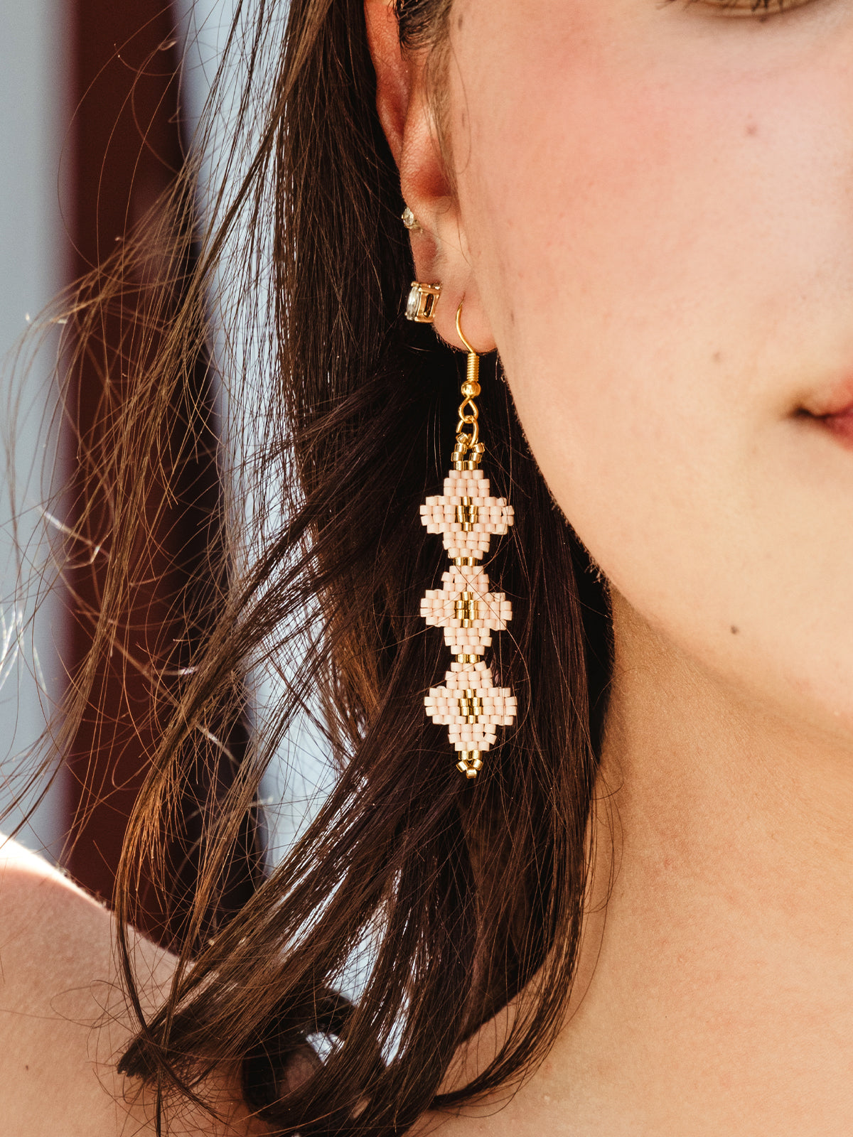 Close-up of a woman's ear showcasing a pair of dangling beaded earrings in soft pink and gold tones. The earrings feature a geometric design made from small beads, capturing the details of the craftsmanship. The background includes strands of the woman's hair and part of a red barn wall.