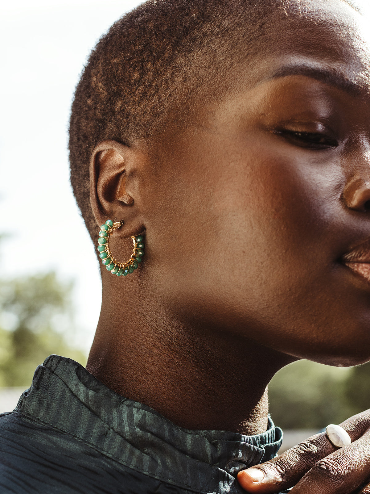 Close-up of a woman with short hair wearing a green top and showcasing a pair of gold hoop earrings with green beaded accents. Her face is partially visible with her eyes gently closed, exuding a calm and confident expression. Her hand, adorned with a white stone ring, rests softly near her neck, adding an elegant touch to the composition. The background is softly blurred, keeping the focus on the jewelry and her serene expression.