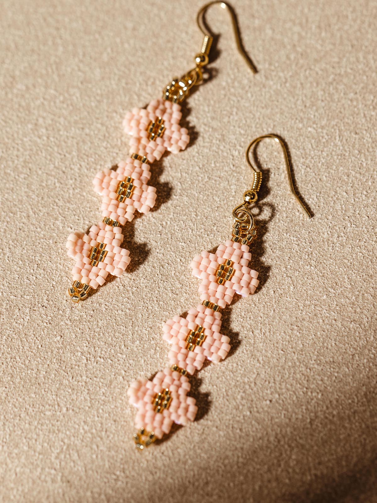 Close-up of handmade dangle earrings with intricate pink and gold beadwork, laid out on a textured beige surface.