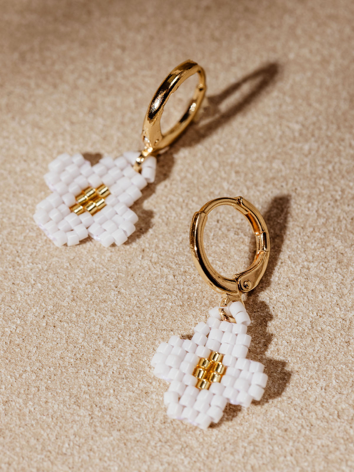 Pair of gold hoop earrings adorned with delicate white and gold beaded flower charms, laid out on a textured beige surface. The intricate beadwork and the elegant gold details create a chic and sophisticated look.