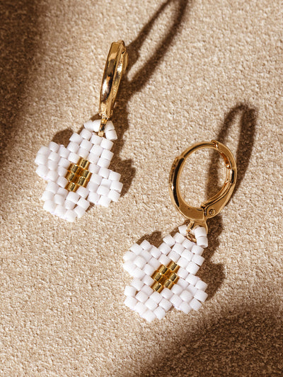 Close-up of gold hoop earrings featuring small beaded flower charms in white and gold, set against a sandy beige background. The earrings' shadows add depth, highlighting the craftsmanship of the beaded design.