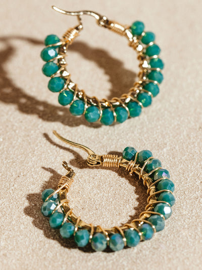 Gold hoop earrings adorned with bright green faceted beads, creating a striking contrast against the gold wire wrapping. The earrings are set on a beige surface, highlighting their elegant and handcrafted design.