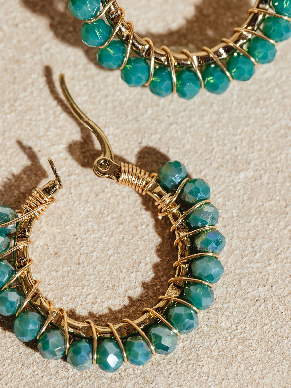 Detailed view of gold hoop earrings with vibrant green faceted beads, meticulously wrapped in gold wire. The earrings are placed on a sandy backdrop, showcasing their luxurious and intricate design.