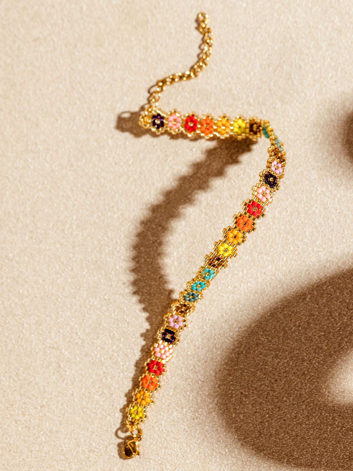 Colorful handmade bracelet featuring intricate beadwork with floral patterns in a rainbow of colors, laid out on a textured beige surface. The delicate craftsmanship is highlighted by the soft shadows and warm lighting.