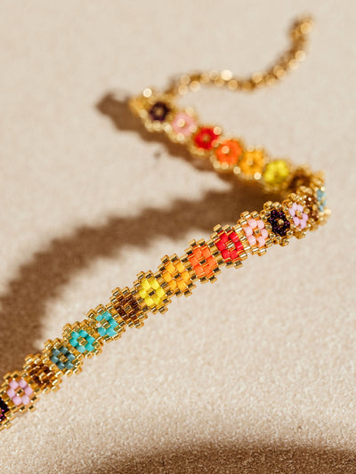 Close-up of a vibrant beaded bracelet showcasing detailed floral patterns in an array of colors, set against a neutral beige background. The bracelet's golden clasp and chain add a touch of elegance to the intricate design.